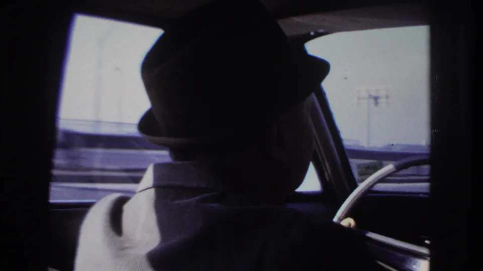 1966 home-movie view from inside a car, showing the driver’s hat and shoulder against a bright Hoboken roadside window.