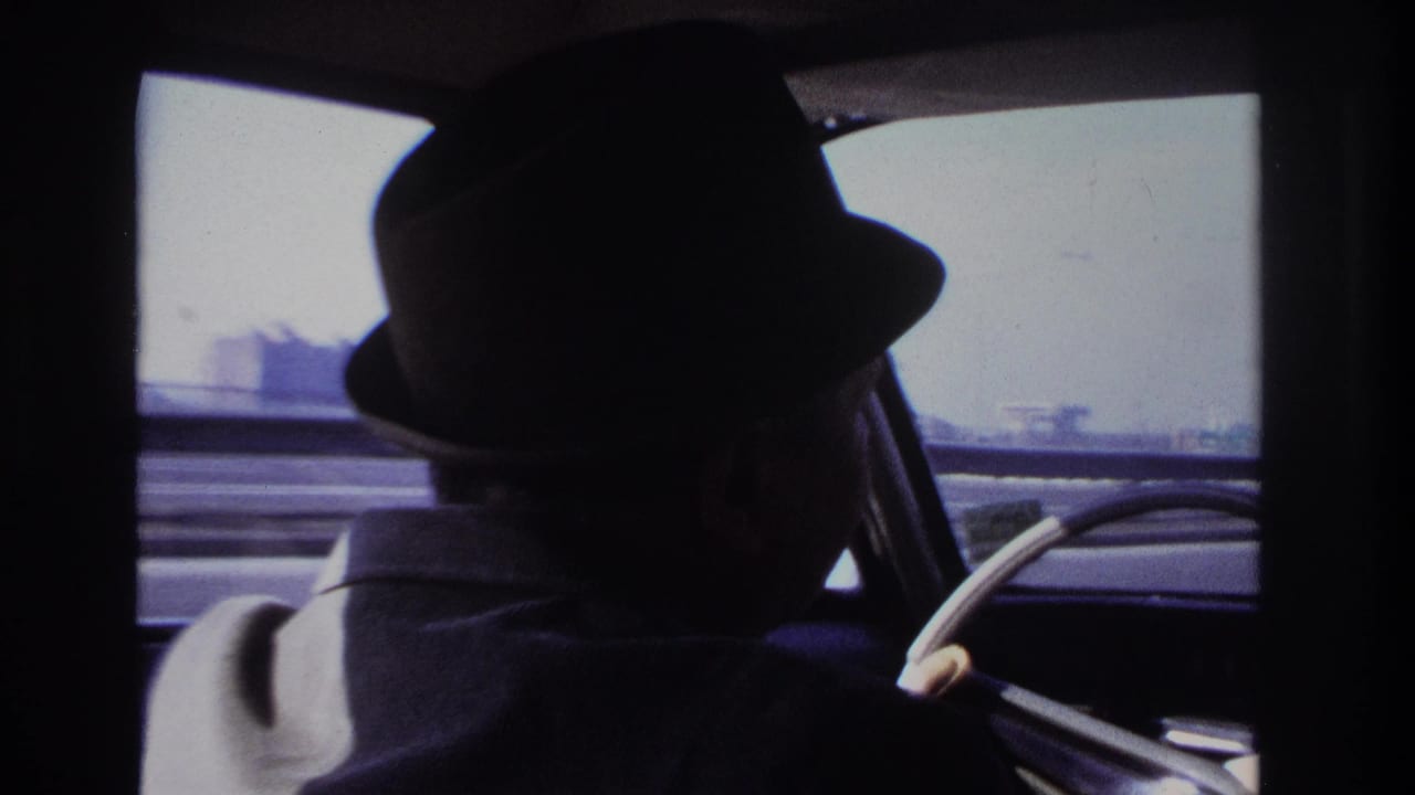 Man in a dark fedora seen from the back seat of a moving car.