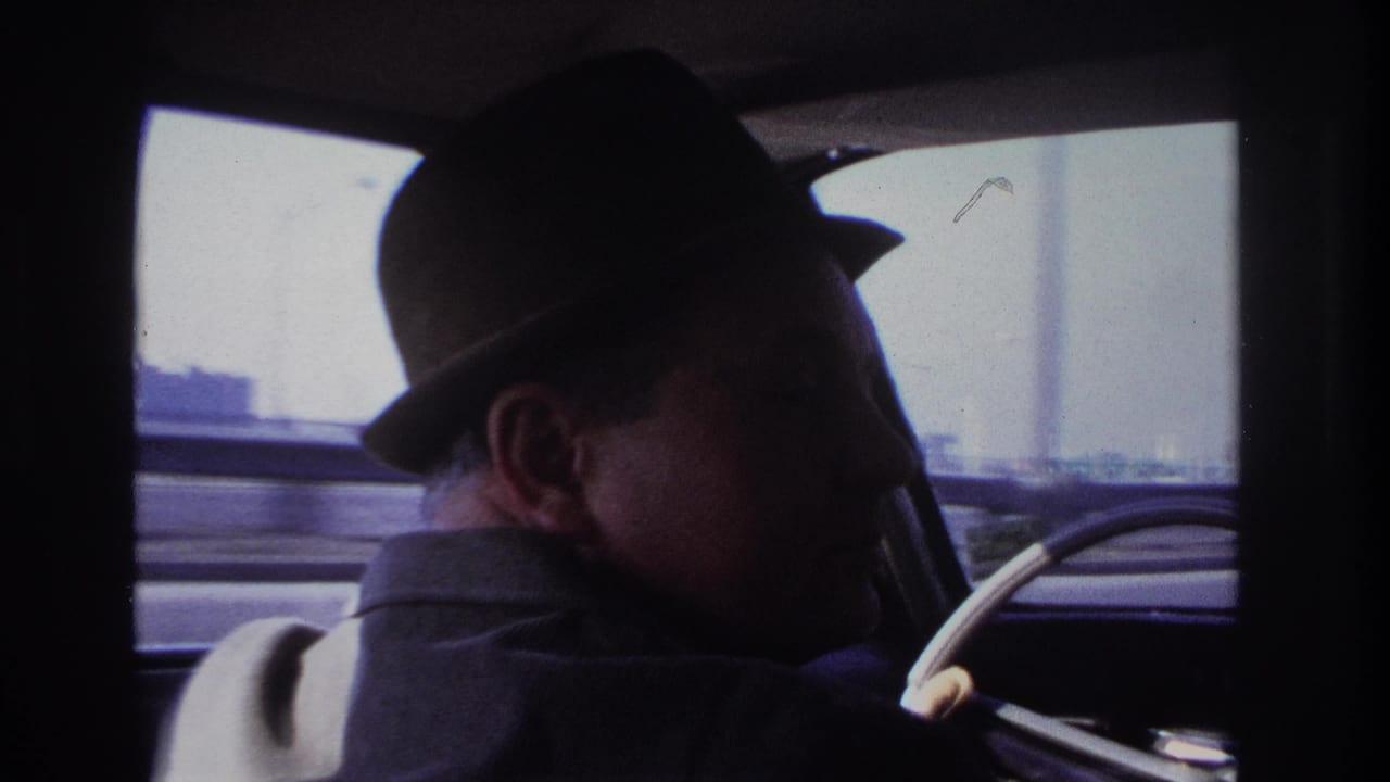 1966 Hoboken home-movie frame with the fedora-wearing driver seen in profile from the rear passenger seat, with a pale window view and steering wheel in front.