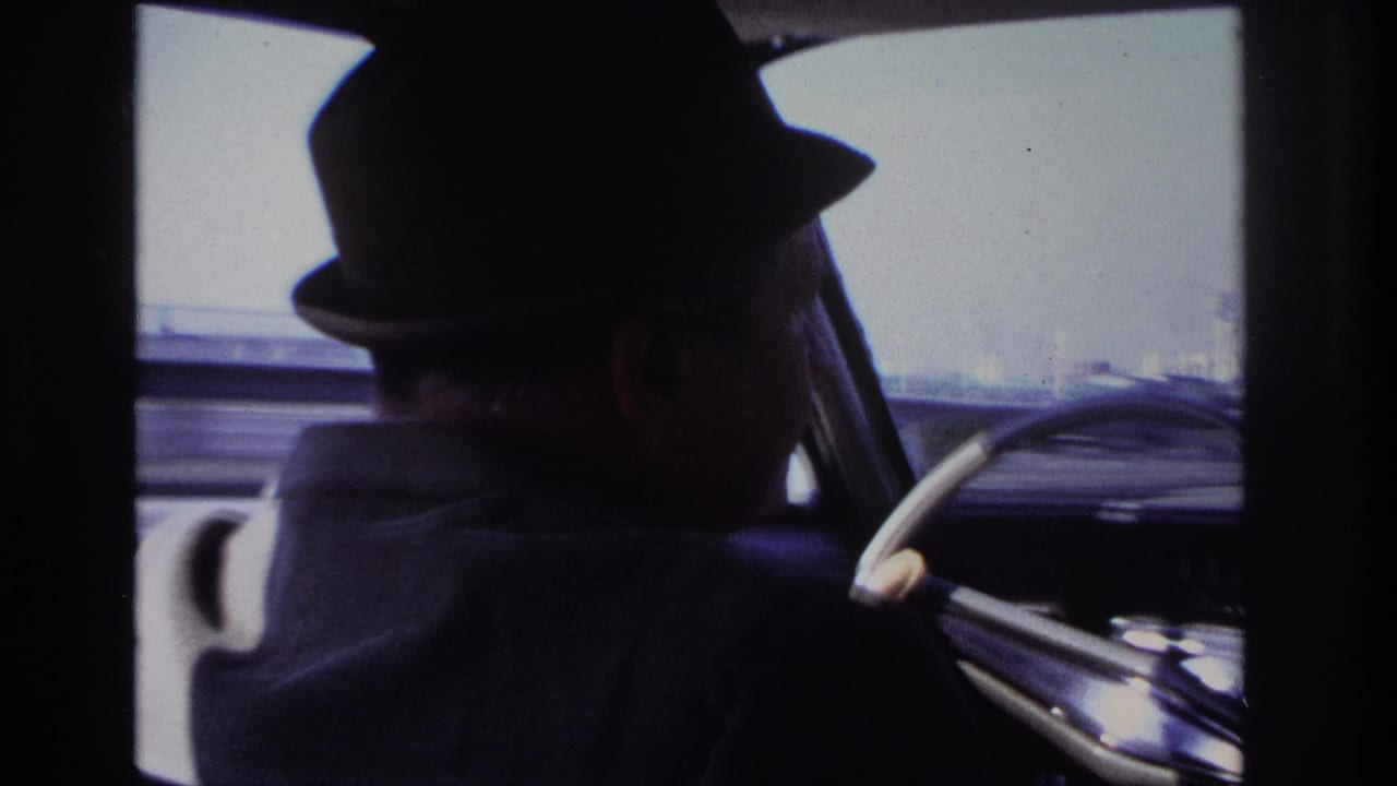 1966 Hoboken home-movie frame showing the driver mostly in silhouette from the back seat, with the steering wheel and blurred exterior roadway visible ahead.