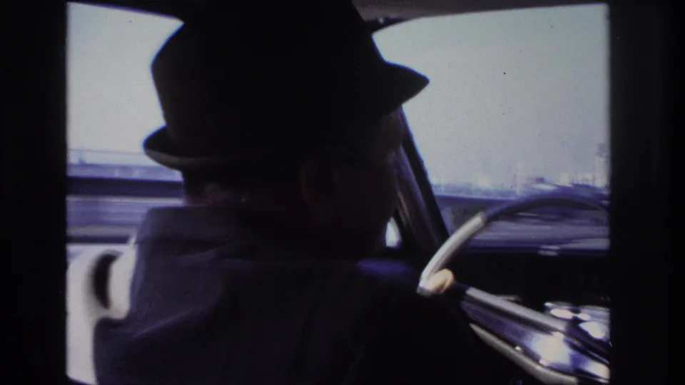 1966 Hoboken home-movie frame showing the driver mostly in silhouette from the back seat, with the steering wheel and blurred exterior roadway visible ahead.