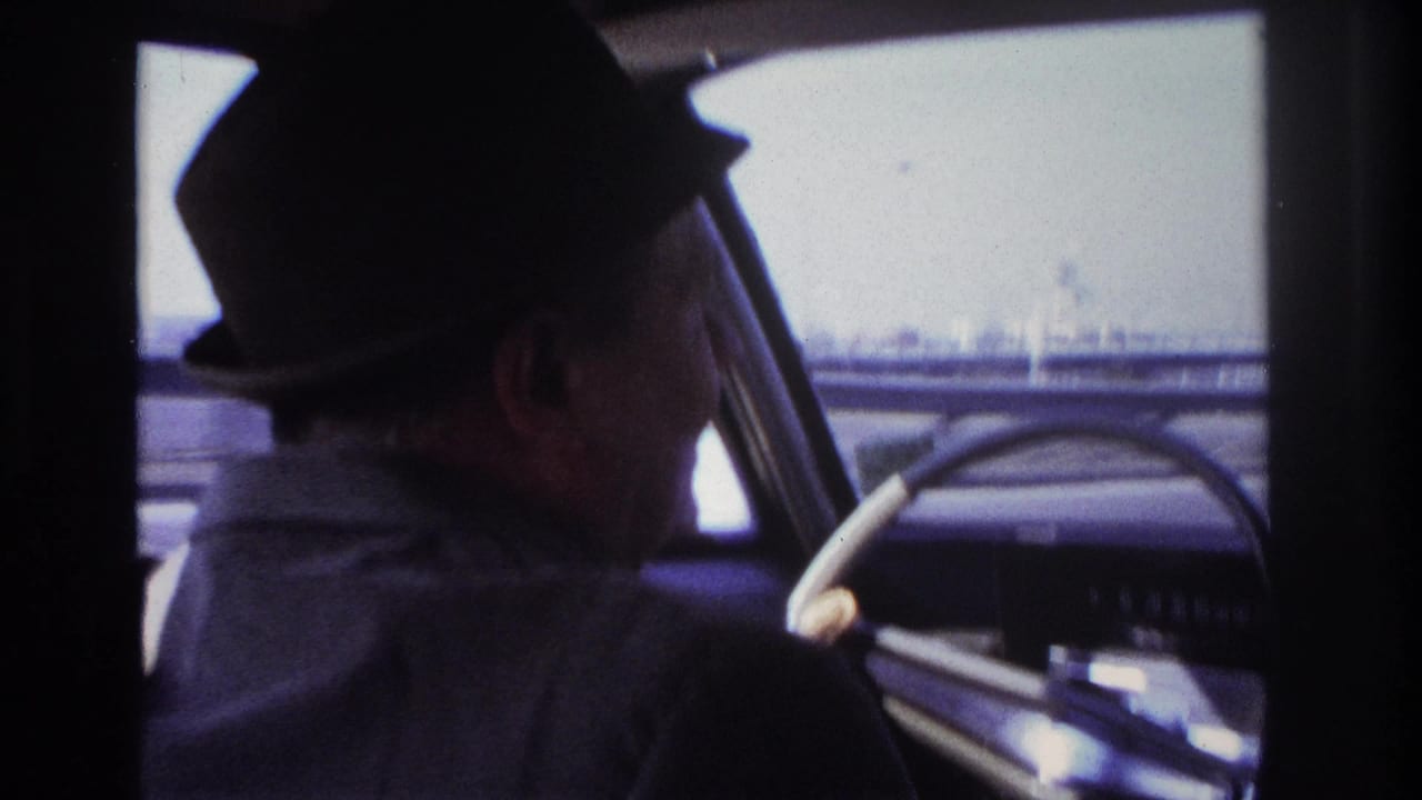 1966 Hoboken home-movie frame of a man in a dark fedora driving a classic car, seen from the back seat with an industrial-looking roadway outside.