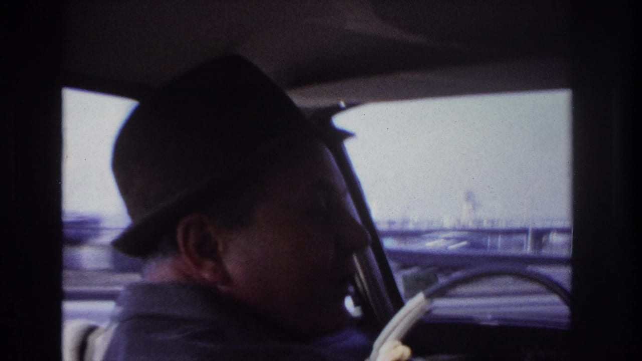 1966 Hoboken home-movie frame of a suited man in a dark fedora driving, viewed from behind inside a classic car.