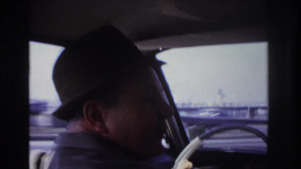 1966 Hoboken home-movie frame of a suited man in a dark fedora driving, viewed from behind inside a classic car.