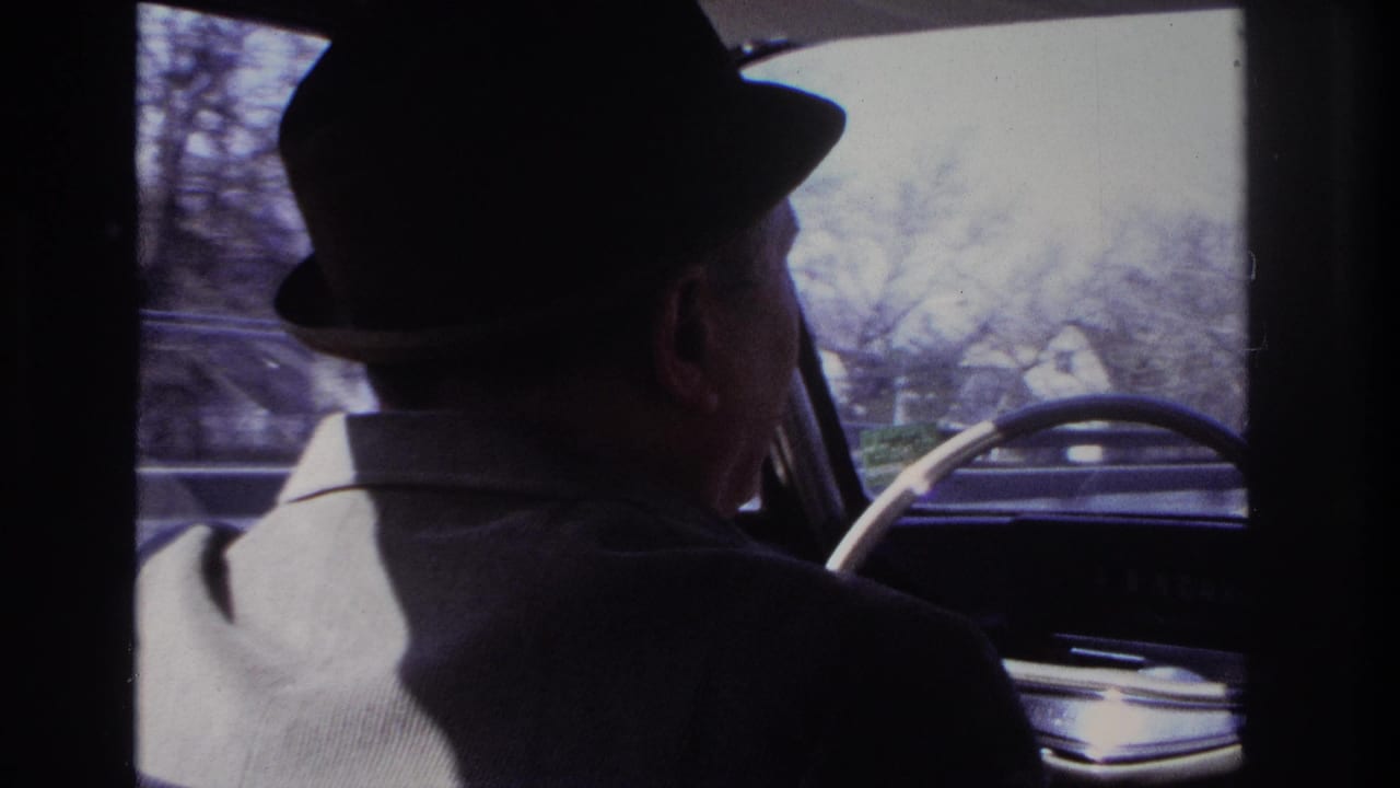 1966 Hoboken-area home-movie frame from the backseat, showing a suited driver and street scene beyond the car windshield.