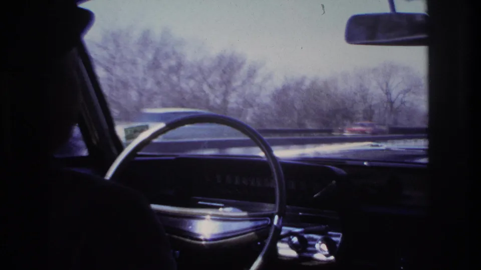 Interior view of a 1960s car in motion near Hoboken, with the driver in silhouette and highway traffic passing outside.