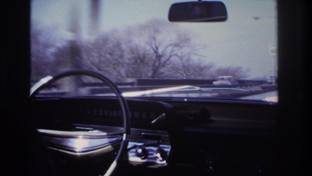 Passenger-seat 8mm view of a classic car dashboard and steering wheel, with winter trees and traffic seen through the windshield.