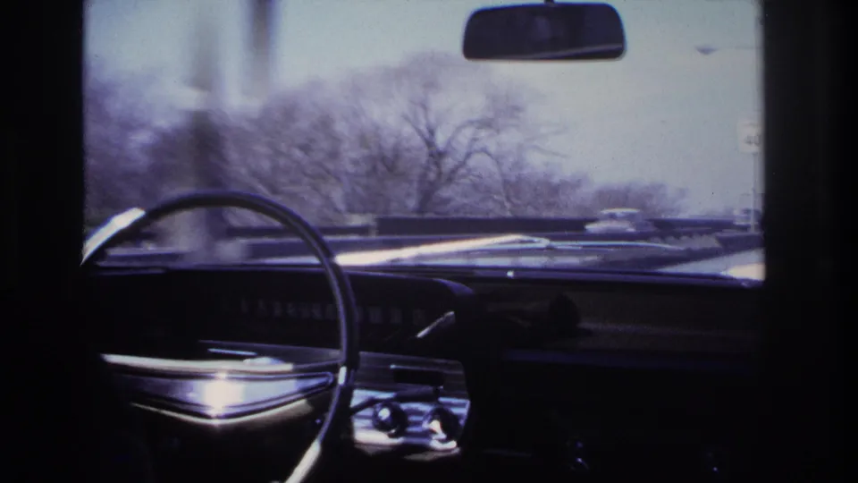 Passenger-seat 8mm view of a classic car dashboard and steering wheel, with winter trees and traffic seen through the windshield.