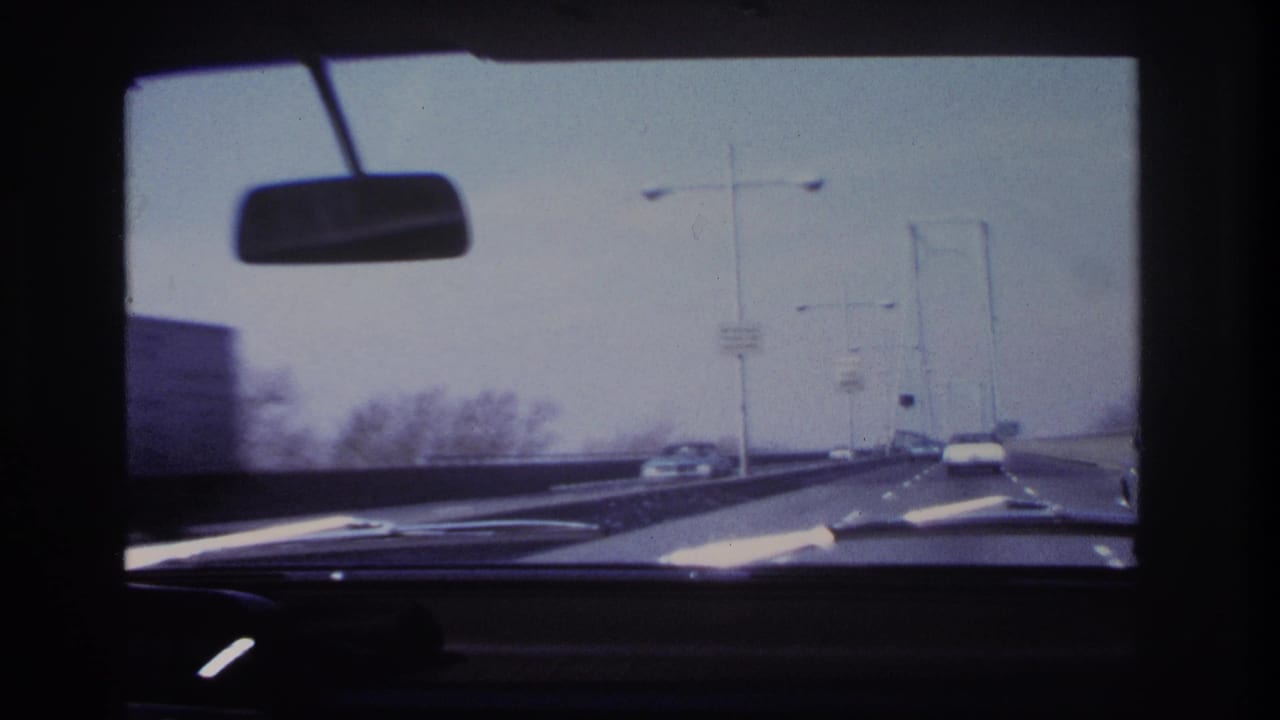 1966 home-movie windshield view from a car near Hoboken, showing a bridge roadway, rearview mirror, dashboard, streetlights, and cars ahead.