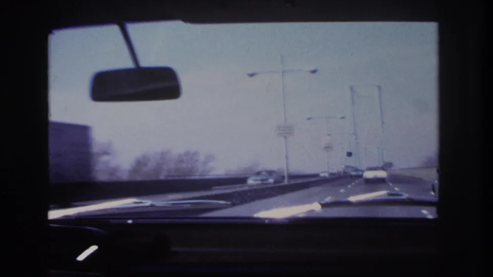 1966 home-movie windshield view from a car near Hoboken, showing a bridge roadway, rearview mirror, dashboard, streetlights, and cars ahead.