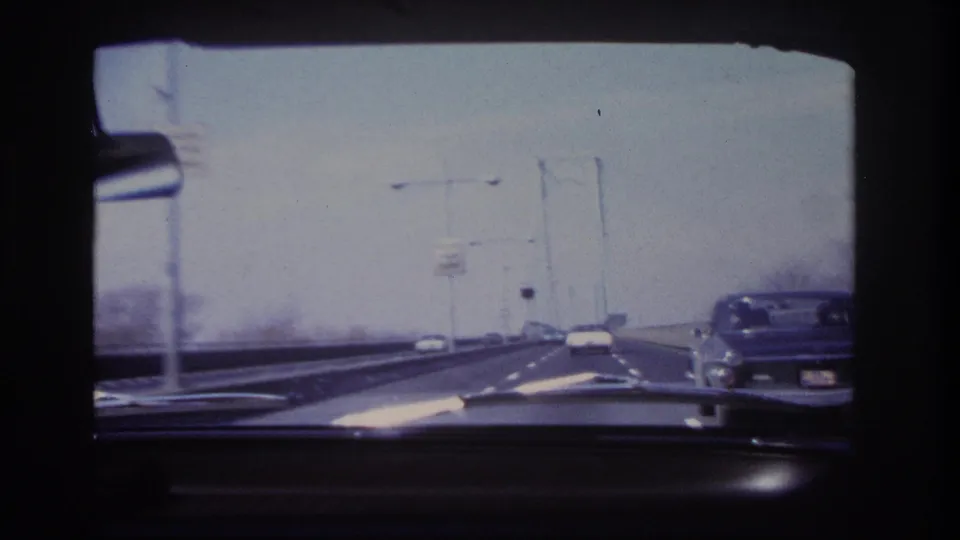 1966 in-car bridge footage near Hoboken, with a dashboard foreground, roadway lights, cars ahead, and a classic car visible along the right edge.