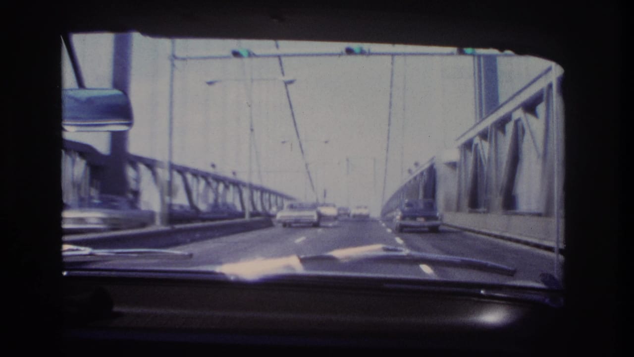 1966 8mm view from inside a moving car on a Hoboken-area bridge, showing steel trusses, suspension cables, dashboard, mirror, and cars ahead.