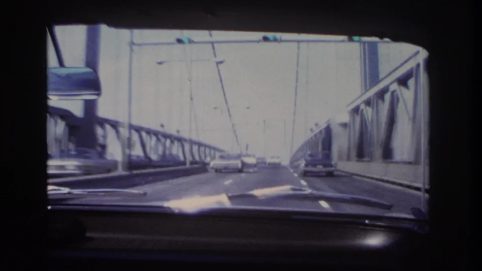1966 8mm view from inside a moving car on a Hoboken-area bridge, showing steel trusses, suspension cables, dashboard, mirror, and cars ahead.