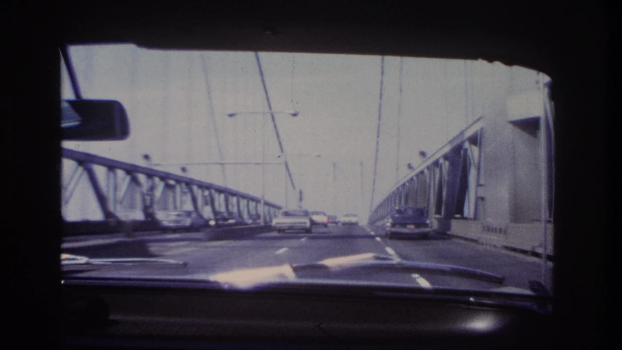 1966 home-movie windshield view on a Hoboken-area bridge, with steel trusses, overhead cables, streetlights, dashboard, and traffic ahead.