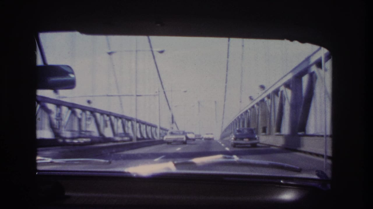1966 8mm view from inside a car crossing a steel suspension bridge near Hoboken, with roadway traffic and cables visible ahead.