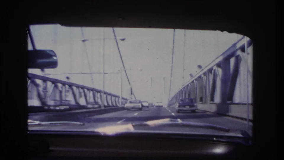 1966 8mm view from inside a car crossing a steel suspension bridge near Hoboken, with roadway traffic and cables visible ahead.