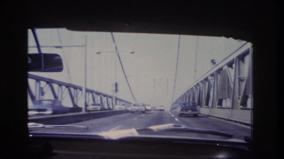 Dashboard-level 8mm frame showing traffic on a suspension bridge in the supplied Hoboken, New Jersey 1966 context.