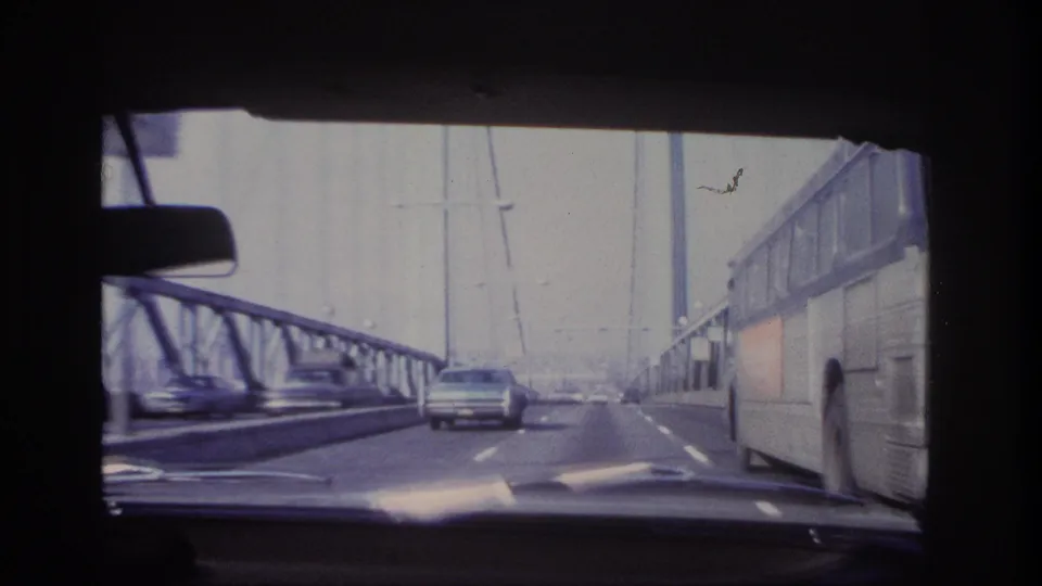 1966 home-movie frame from a moving car as a large coach bus passes on the right side of a steel bridge roadway.