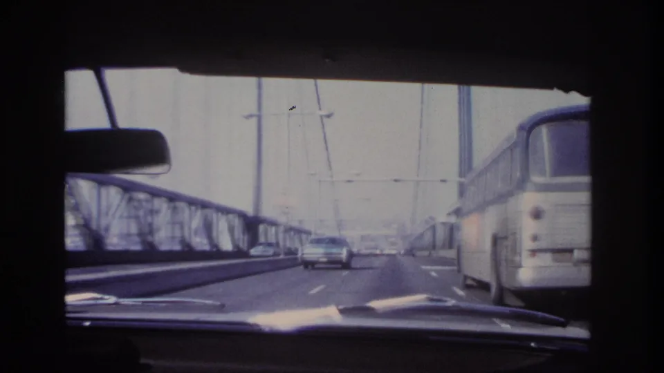 1966 Hoboken-area bridge crossing seen from inside a car, with a bus to the right, a sedan ahead, steel railings, and suspension cables.