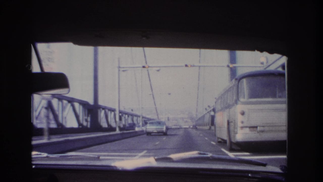 Bridge roadway seen through a windshield with a bus close at right and a car ahead.