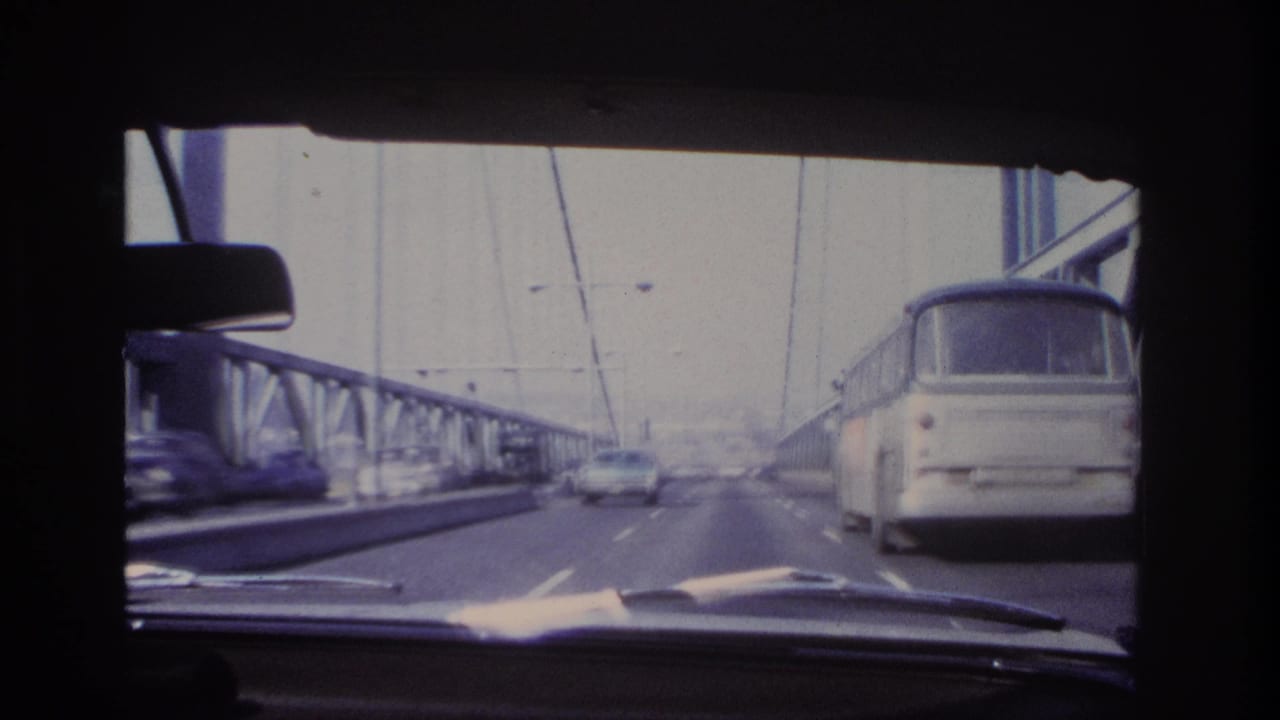 From inside a moving car, 1966 bridge traffic appears with a bus in the right lane, a car ahead, railings, cables, and faded 8mm color.