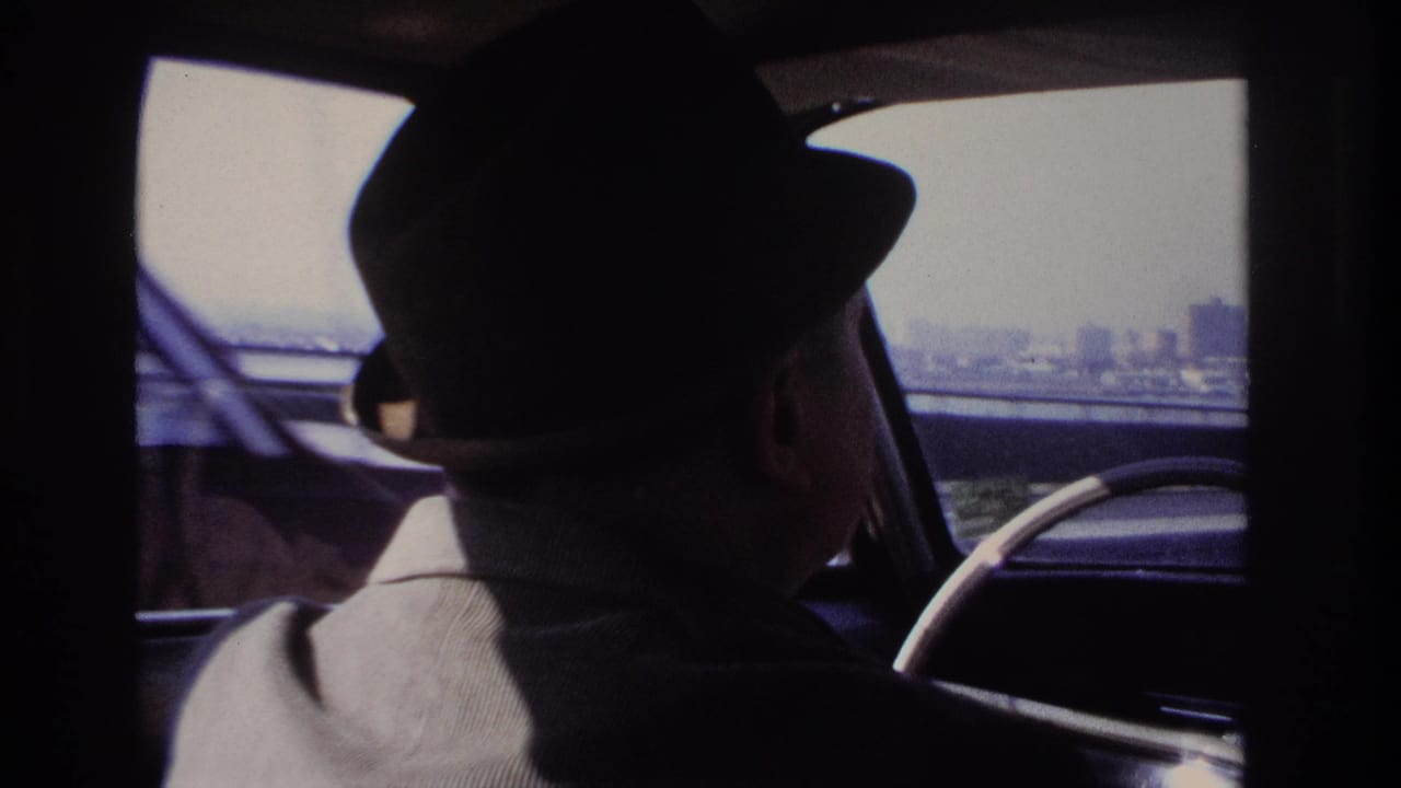 Inside a car near Hoboken in 1966, a driver in a fedora is seen from behind with bridge structure and distant skyline beyond.