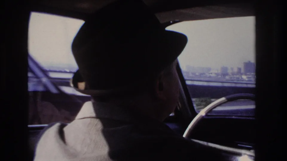 Inside a car near Hoboken in 1966, a driver in a fedora is seen from behind with bridge structure and distant skyline beyond.