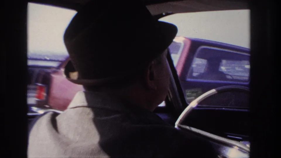 1966 Hoboken-area home movie frame showing an older man in a fedora driving, with passing cars visible through the window.
