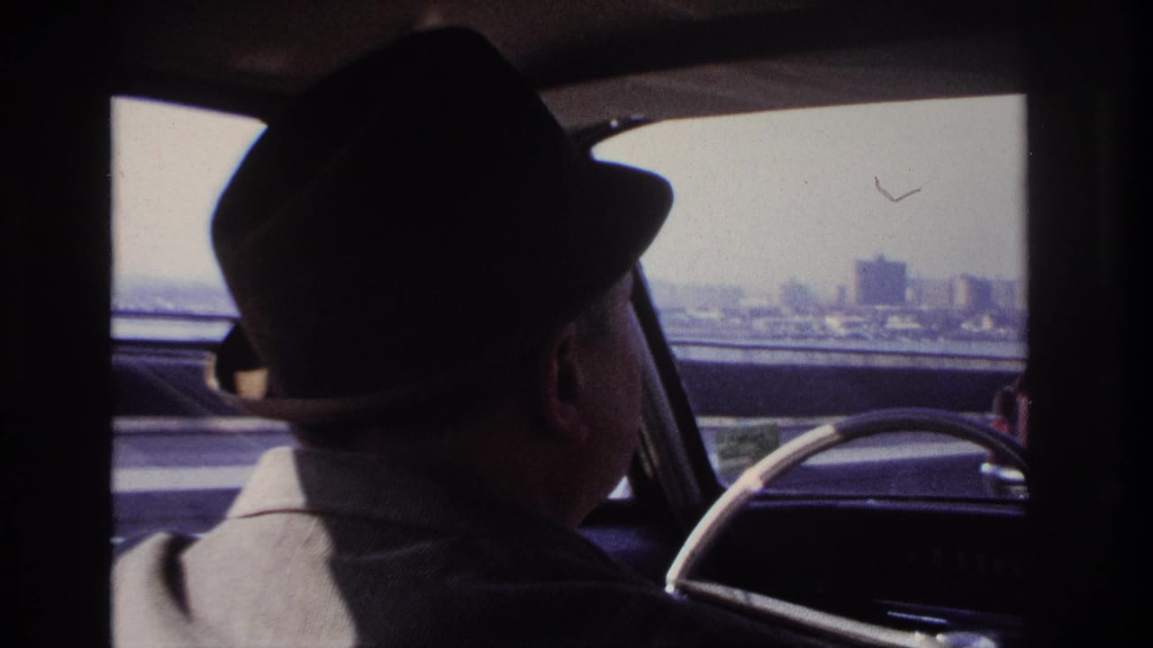 1966 Hoboken-area car interior frame showing the driver and a distant city skyline beyond the roadside.