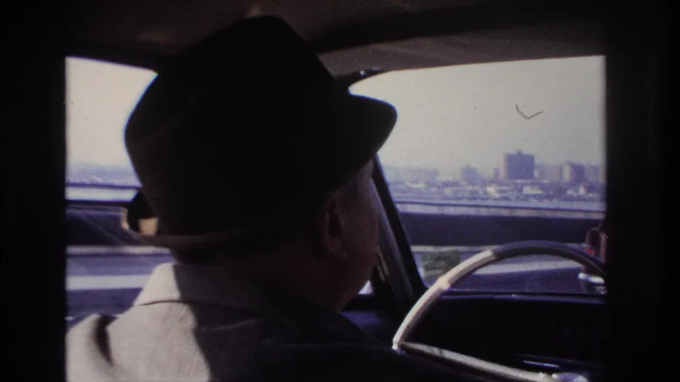 1966 Hoboken-area car interior frame showing the driver and a distant city skyline beyond the roadside.