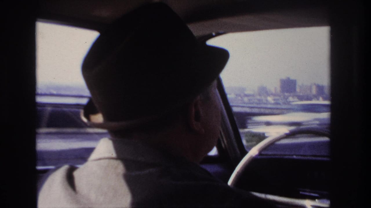 Final 1966 Hoboken-area frame with the driver in shadow, the steering wheel at right, and tall buildings in the distance.
