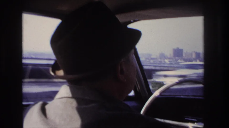 Final 1966 Hoboken-area frame with the driver in shadow, the steering wheel at right, and tall buildings in the distance.