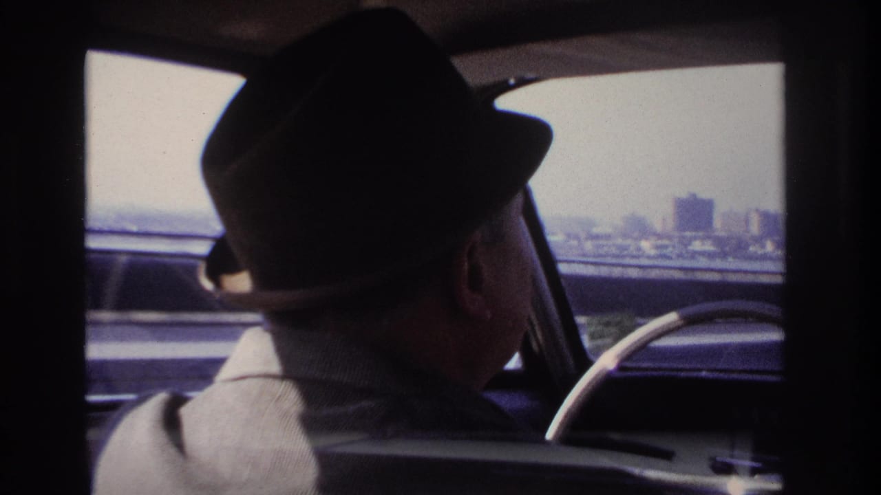 1966 Hoboken-area 8mm frame showing a fedora-wearing driver inside a car, with freeway traffic and distant urban structures outside.