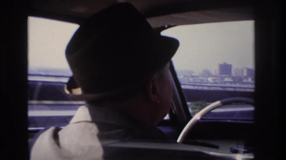 1966 Hoboken-area 8mm frame showing a fedora-wearing driver inside a car, with freeway traffic and distant urban structures outside.