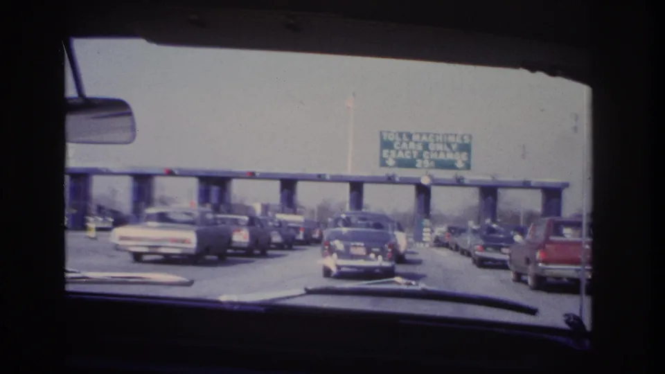 From inside a car, mid-1960s traffic approaches toll machines beneath a green sign for exact change cars only.