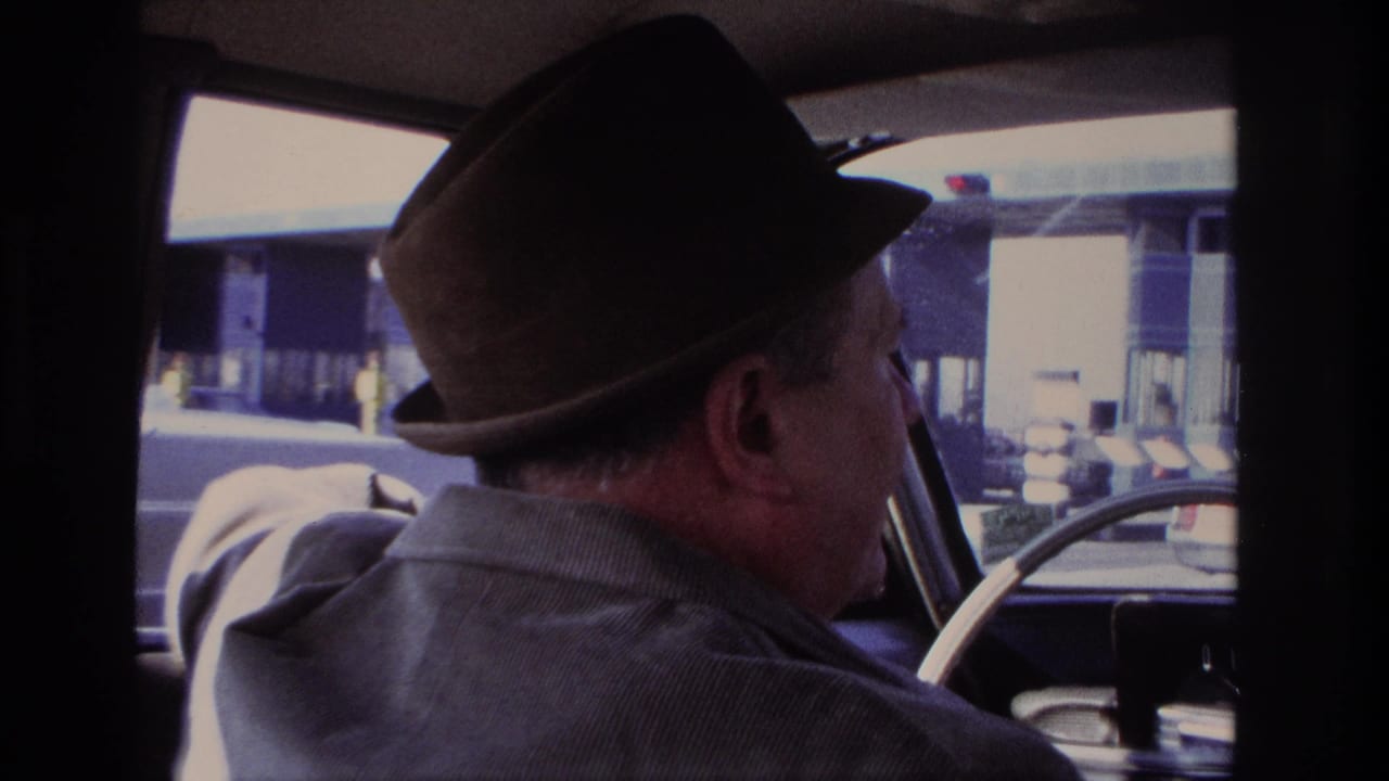 A man in a brown fedora and corduroy jacket is seen in profile driving near toll booth lanes in the 1966 home movie.