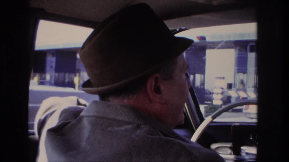 A man in a brown fedora and corduroy jacket is seen in profile driving near toll booth lanes in the 1966 home movie.