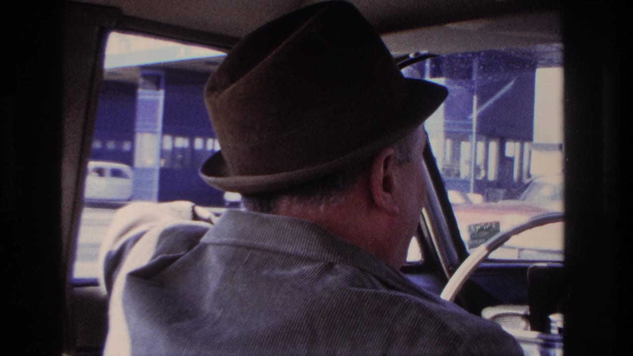 Older man in a brown fedora and gray jacket drives through Hoboken in 1966, viewed from inside the rear seat of the car.