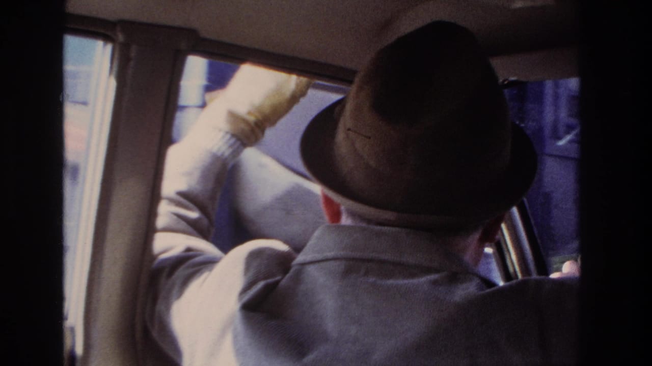 1966 Hoboken home-movie frame showing a man in a brown fedora and gray coat from behind as he sits at the wheel.