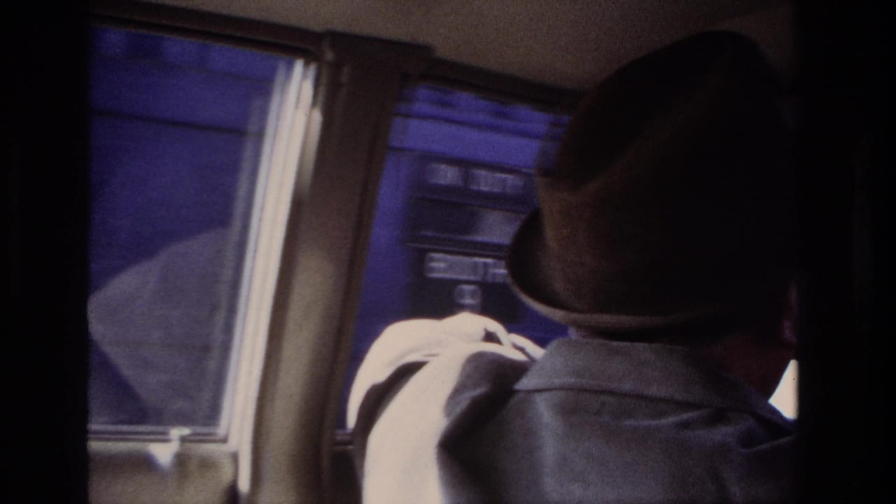 Driver in fedora near a car window with another vehicle or dark exterior surface outside.