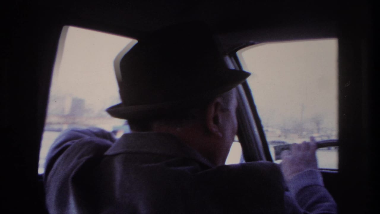 Home-movie frame of the Hoboken driver in a fedora steering past bright exterior buildings and industrial forms.