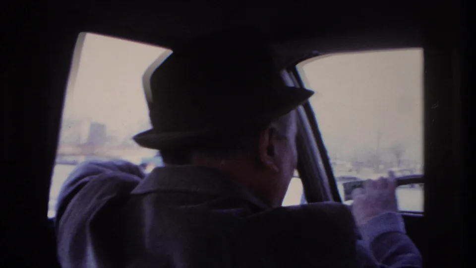 Home-movie frame of the Hoboken driver in a fedora steering past bright exterior buildings and industrial forms.