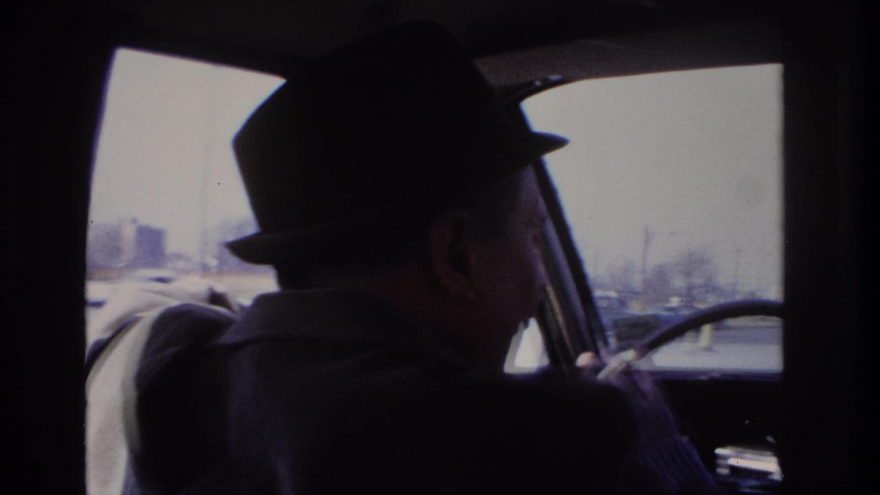 1966 Hoboken home-movie frame showing a man in a dark fedora and coat at the wheel, with traffic visible through the windshield.