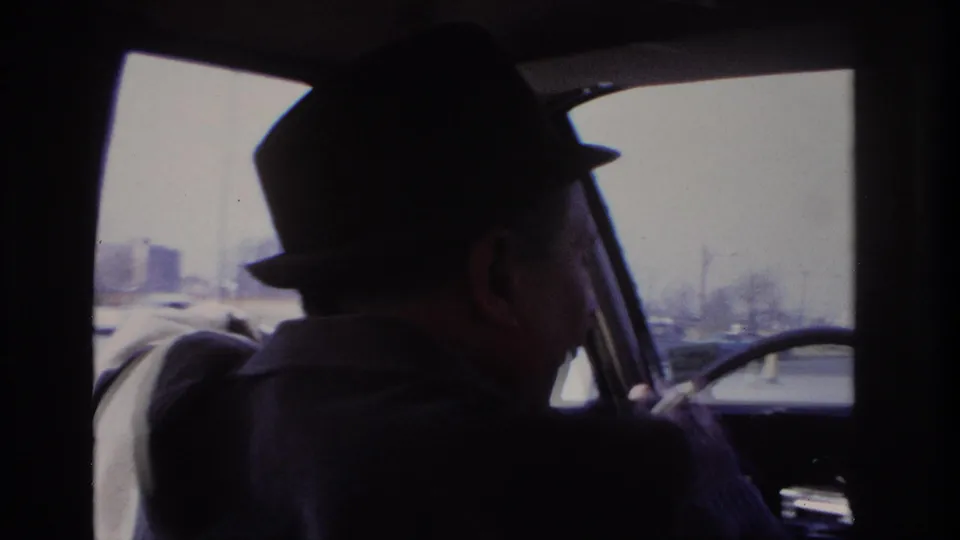 1966 Hoboken home-movie frame showing a man in a dark fedora and coat at the wheel, with traffic visible through the windshield.