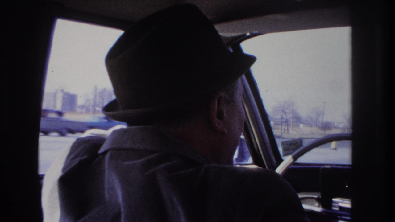Driver in fedora looks ahead through the windshield of a car.