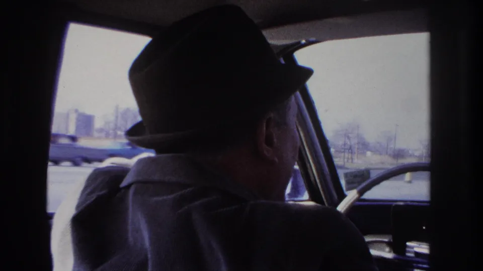 1966 Hoboken footage frame from inside a car, showing the driver in a fedora with a blue car passing outside.