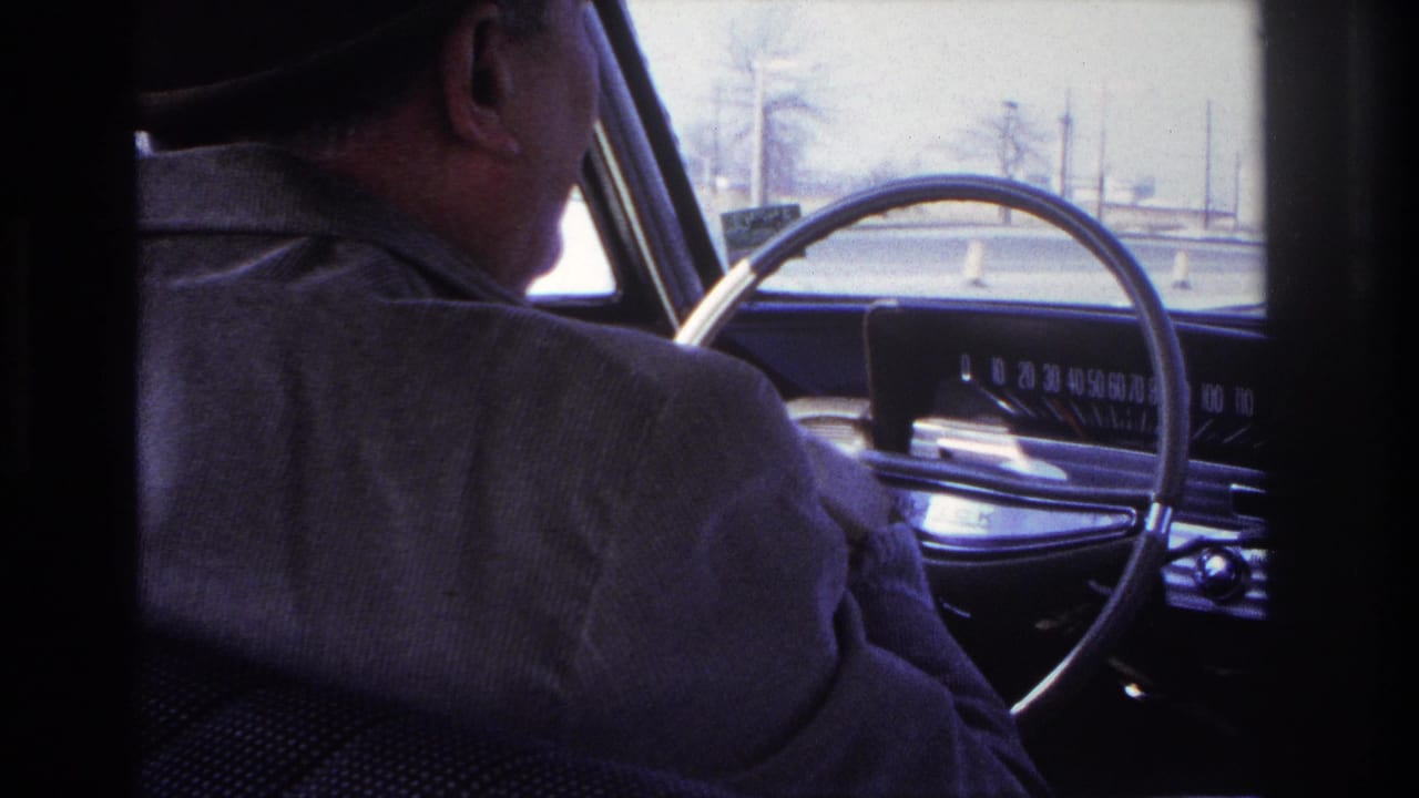 1966 Hoboken car-interior frame showing a fedora-wearing driver beside a large steering wheel, dashboard, windshield, and traffic cones ahead.