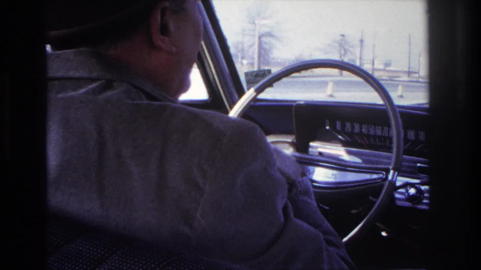 1966 Hoboken car-interior frame showing a fedora-wearing driver beside a large steering wheel, dashboard, windshield, and traffic cones ahead.