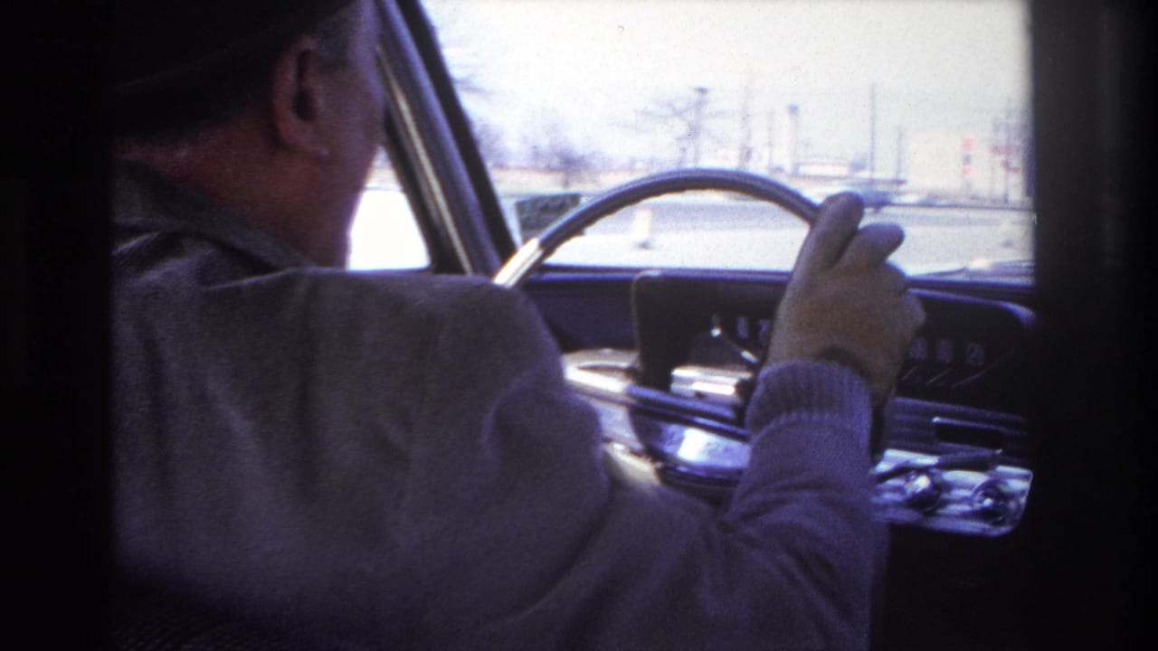 1966 Hoboken home-movie frame showing an older man in a fedora and gloves driving a mid-century car from the passenger-side view.