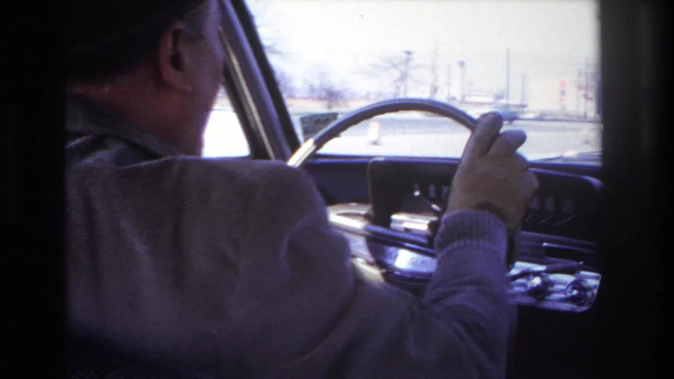 1966 Hoboken home-movie frame showing an older man in a fedora and gloves driving a mid-century car from the passenger-side view.